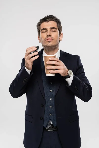Hombre de negocios con los ojos cerrados sosteniendo taza de papel y tapa mientras huele café de la mañana aislado en gris - foto de stock