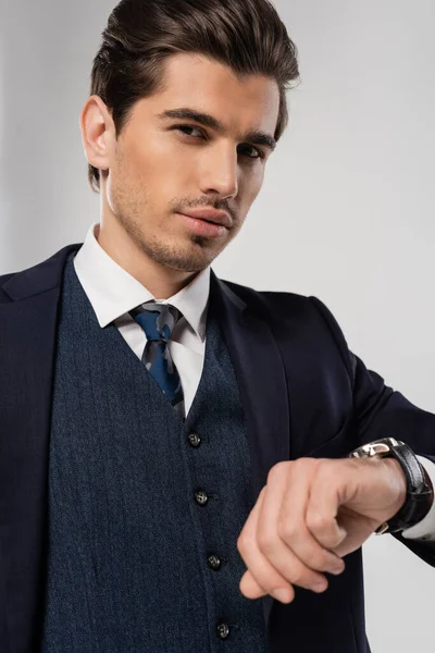 Молодий бізнесмен з наручним годинником стоїть в костюмі і дивиться на камеру ізольовано на сірому — стокове фото