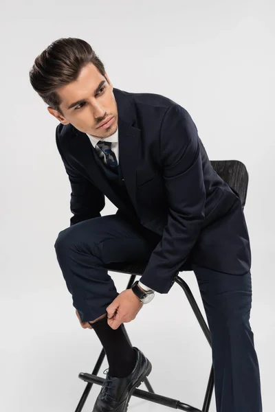 Joven hombre de negocios en ropa formal sentado en la silla mientras se ajusta calcetín aislado en gris - foto de stock
