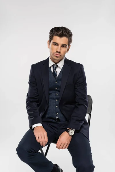 Joven hombre de negocios en ropa formal sentado en la silla mientras mira la cámara aislada en gris - foto de stock
