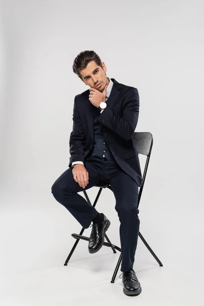 Повна довжина молодого бізнесмена в офіційному одязі, сидячи на стільці, позує на сірому — стокове фото