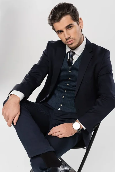 Uomo d'affari fiducioso in abbigliamento formale seduto sulla sedia mentre posa isolato sul grigio — Foto stock