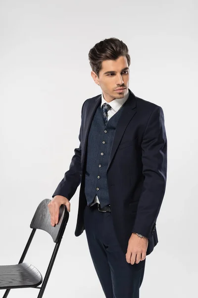 Молодой бизнесмен в формальной одежде, стоящий рядом со стулом, позируя изолированным на серой — стоковое фото