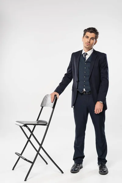 Повна довжина молодого бізнесмена в офіційному одязі, стоячи біля стільця, позуючи на сірому — стокове фото