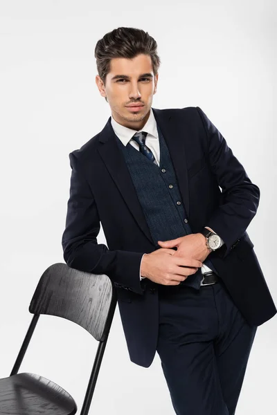 Giovane uomo d'affari in abito formale in piedi vicino alla sedia mentre guarda la fotocamera isolata sul grigio — Foto stock