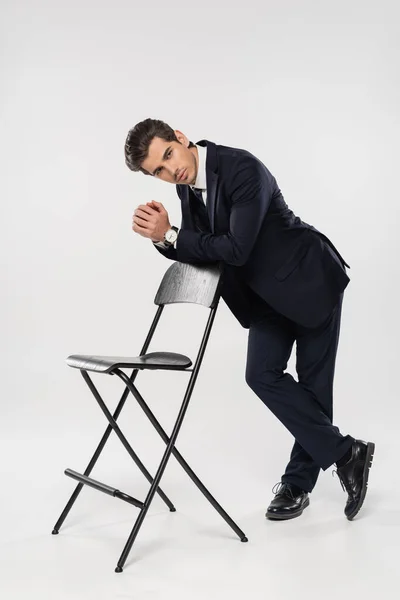 Comprimento total do empresário bem sucedido no desgaste formal em pé perto da cadeira no cinza — Fotografia de Stock