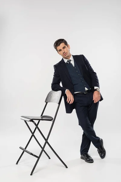 Longitud completa de exitoso hombre de negocios en traje con chaqueta de pie cerca de la silla en gris - foto de stock