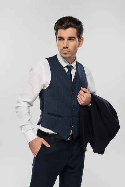 Hombre de negocios en ropa formal sosteniendo blazer de pie con la mano en el bolsillo aislado en gris - foto de stock