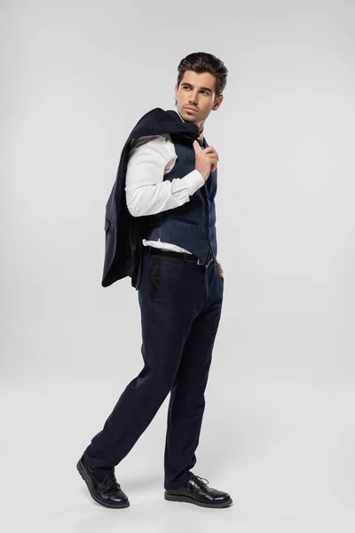 Полная длина бизнесмена в формальной одежде, держащего блейзер и стоящего с рукой в кармане на сером — стоковое фото