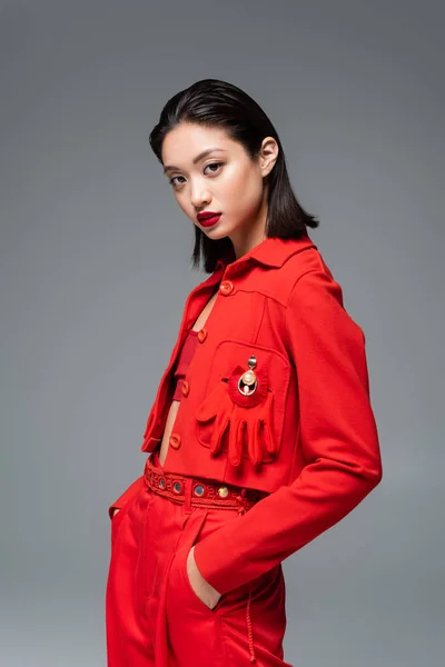 Азиатка в красной куртке, украшенной брошь и перчаткой, стоящая с руками в карманах, изолированных на сером — стоковое фото