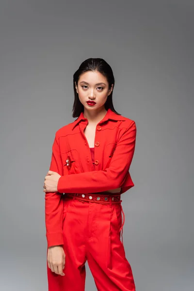 Брюнетка азіатська жінка в червоному модному костюмі дивиться на камеру ізольовано на сірому — стокове фото