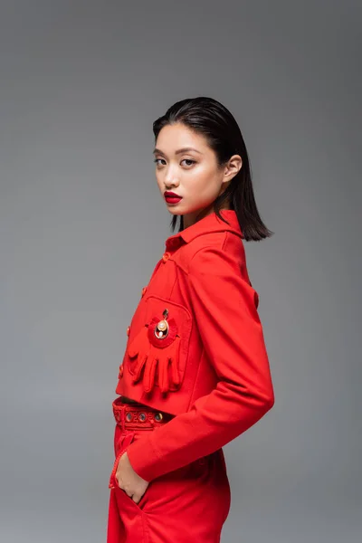 Modelo asiático morena en chaqueta roja decorada con guante posando con mano en bolsillo aislado en gris - foto de stock