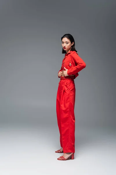 In voller Länge junge asiatische Frau in roter Jacke und Hose schaut in die Kamera auf grauem Hintergrund — Stockfoto