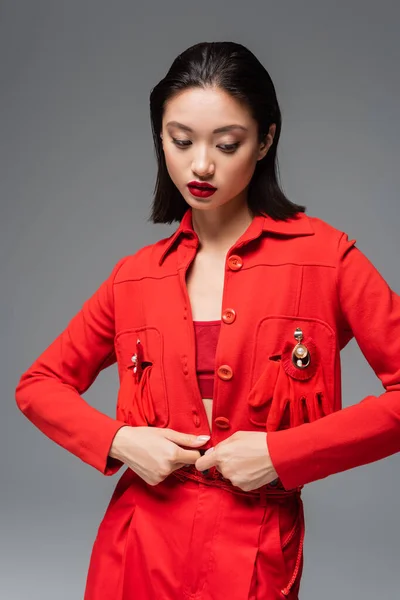 Молода азіатка в червоній куртці, прикрашена брошками та рукавичками, ізольована на сірому — стокове фото