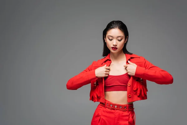 Brunette asian woman touching red fashionable jacket isolated on grey — Stock Photo