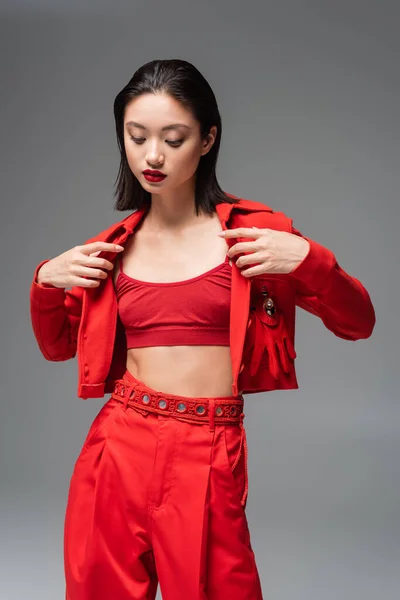 Brunette asian woman in top and pants taking off red jacket isolated on grey — Stock Photo