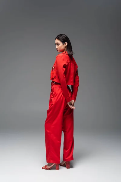 Piena lunghezza di donna asiatica in giacca rossa e pantaloni in piedi su sfondo grigio — Foto stock