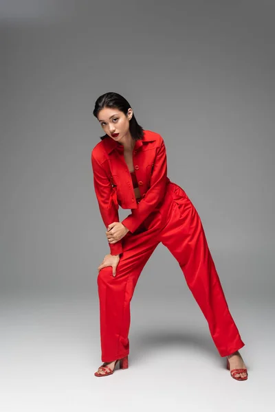 Longitud completa de la elegante mujer asiática en chaqueta roja y pantalones mirando a la cámara mientras posando sobre fondo gris - foto de stock