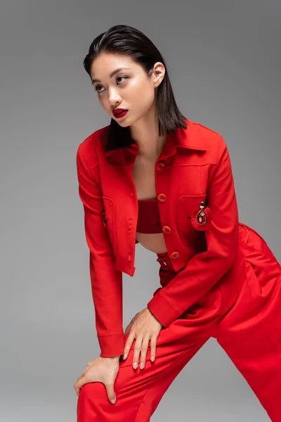 Брюнетка азіатська жінка в червоному елегантному костюмі дивиться вбік, позує ізольовано на сірому — стокове фото
