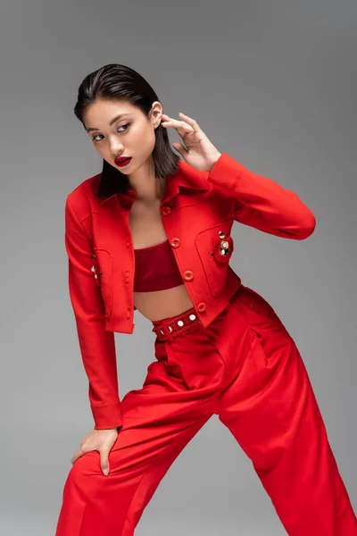 Morena asiática mujer vistiendo rojo elegante chaqueta y pantalones posando aislado en gris - foto de stock