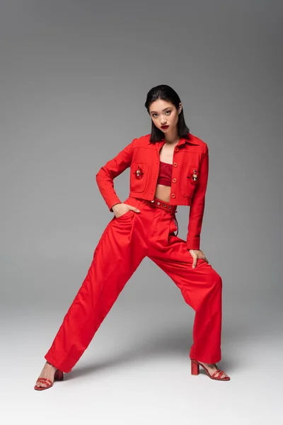 Полная длина азиатской женщины в красном элегантном костюме позирует с рукой на бедре на сером фоне — стоковое фото