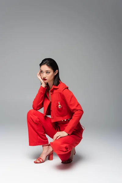 Longitud completa de la mujer asiática en rojo y elegante traje sentado en haunches con la mano cerca de la cara sobre fondo gris - foto de stock