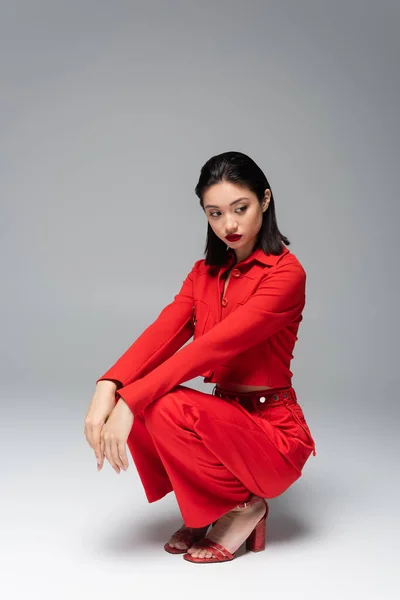 Pretty asian woman in red stylish outfit sitting on haunches on grey background — Stock Photo