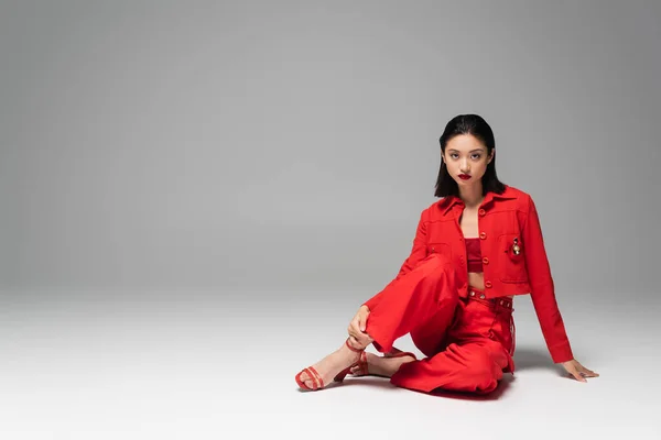 Morena mujer asiática en pantalones rojos y chaqueta sentado sobre fondo gris - foto de stock