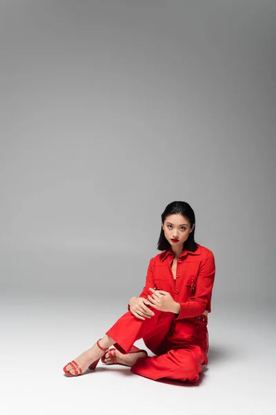 Brunetta modello asiatico in giacca rossa e pantaloni guardando la fotocamera mentre seduto su sfondo grigio — Foto stock