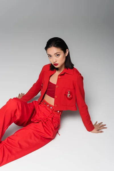 Молодая азиатка в красном модном костюме смотрит в камеру и сидит на сером фоне — стоковое фото