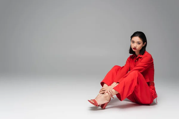 Junge asiatische Frau blickt in die Kamera, während sie im roten trendigen Outfit auf grauem Hintergrund sitzt — Stockfoto