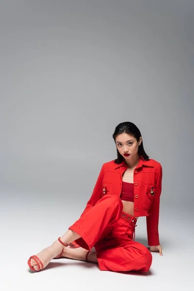 Morena mujer asiática en rojo chaqueta de moda y pantalones sentado y mirando a la cámara sobre fondo gris - foto de stock