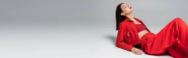 Morena asiática mulher no vermelho elegante roupa posando com olhos fechados no fundo cinza, banner — Fotografia de Stock