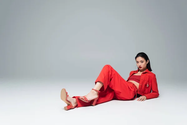 Молодая азиатка в красной стильной одежде и сандалиях на каблуках позирует на сером фоне — стоковое фото