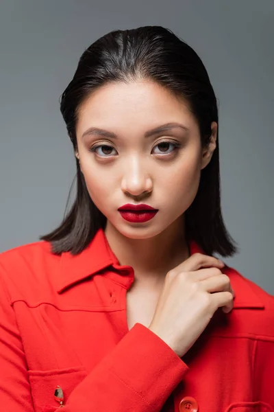 Retrato de morena mujer asiática con labios rojos vistiendo chaqueta roja y mirando a la cámara aislada en gris - foto de stock