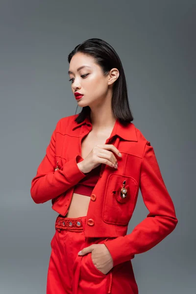 Азиатка в красных брюках и куртке с брошь позирует с рукой в кармане изолированы на сером — стоковое фото