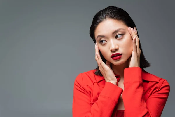 Retrato de mujer asiática de moda con labios rojos tomados de la mano cerca de la cara aislada en gris - foto de stock