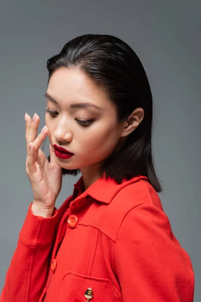 Joven mujer asiática en elegante chaqueta tocando la cara con maquillaje y labios rojos aislados en gris - foto de stock