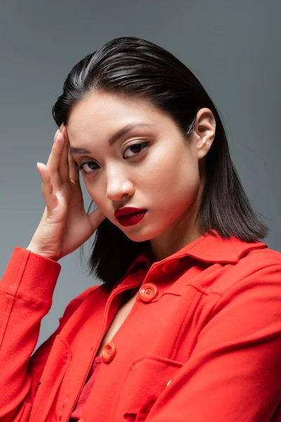 Retrato de mujer asiática con maquillaje y labios rojos usando chaqueta de moda aislada en gris - foto de stock