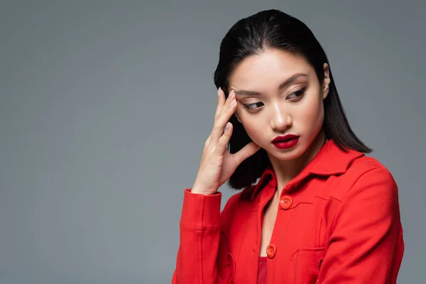 Retrato de morena mujer asiática en chaqueta roja cogida de la mano cerca de la cara y mirando hacia otro lado aislado en gris - foto de stock