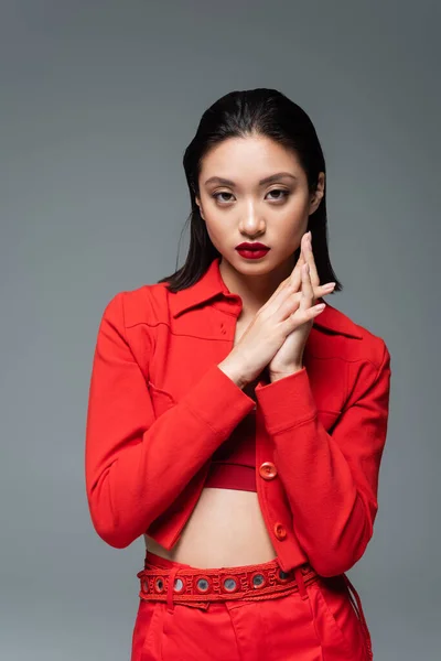 Morena modelo asiático en rojo chaqueta de moda mirando a cámara aislada en gris - foto de stock