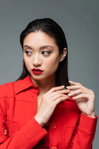 Bonita mujer asiática con maquillaje y labios rojos vistiendo chaqueta de moda y mirando a otro lado aislado en gris - foto de stock