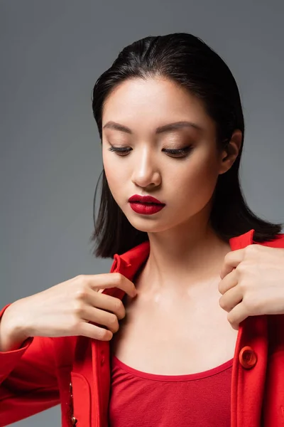 Молодая азиатка с красными губами трогает стильную куртку, изолированную на сером — стоковое фото