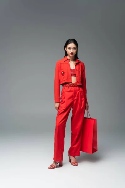 Full length of asian model in red jacket and trousers standing with shopping bag on grey background — Stock Photo