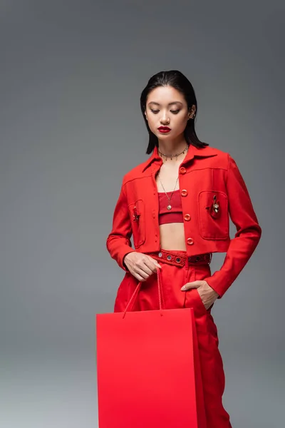 Morena mujer asiática en rojo elegante atuendo sosteniendo bolsa de compras y mano en bolsillo aislado en gris - foto de stock