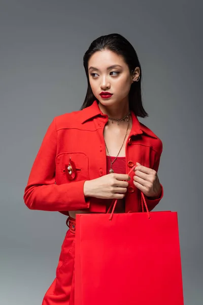 Mujer asiática en chaqueta roja y collares sosteniendo bolsa de compras y mirando hacia otro lado aislado en gris - foto de stock