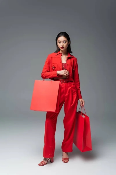 Volle Länge der asiatischen Frau in stylischer Jacke und Hose, die Einkaufstaschen hält, während sie vor grauem Hintergrund wegschaut — Stockfoto