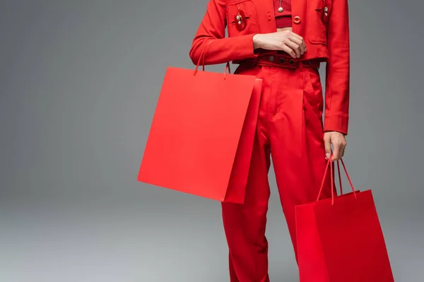 Vista parcial de la mujer en traje rojo de pie con bolsas de compras sobre fondo gris - foto de stock