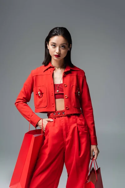 Morena mujer asiática en rojo ropa de moda posando con la mano en el bolsillo y bolsas aisladas en gris - foto de stock
