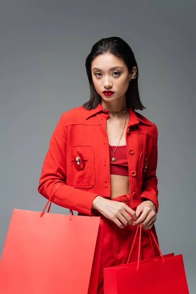 Модная азиатская женщина в красной куртке, держащая сумки с покупками и глядя на камеру, изолированную на сером — стоковое фото